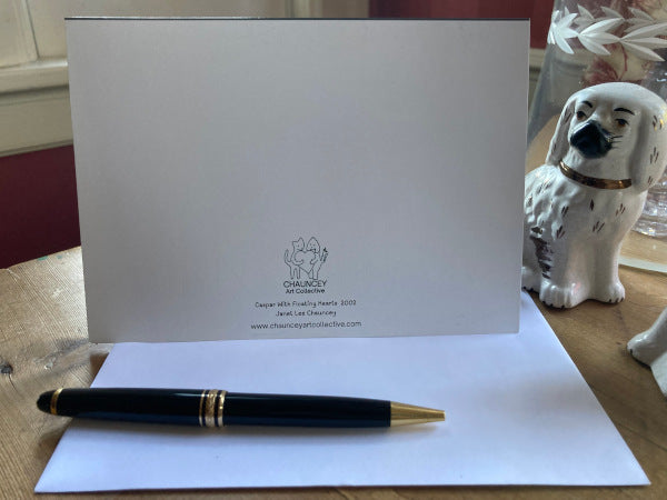 Front of the folded card Caspar With Floating Hearts is displayed on a brown desk with a white envelope, gold pen, and black and white Staffordshire dog statue to the right.