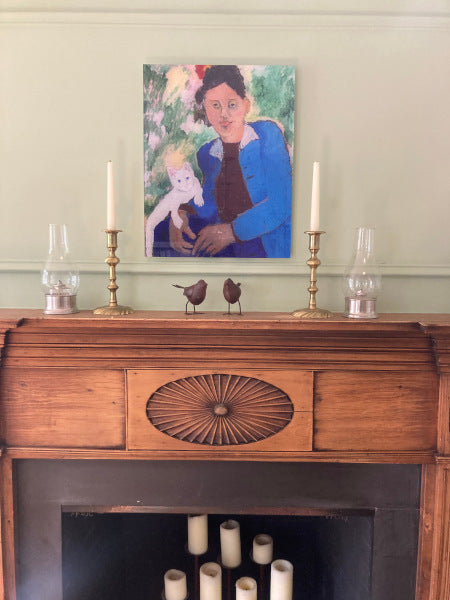 Mockup of Erica Blackwell With Caspar, an acrylic portrait print is seen mounted on the green wall centered above a pine mantle with candlesticks and, hurricanes and two metal bird figurines.  This photo accurately shows the art print in a realistic setting in a home.