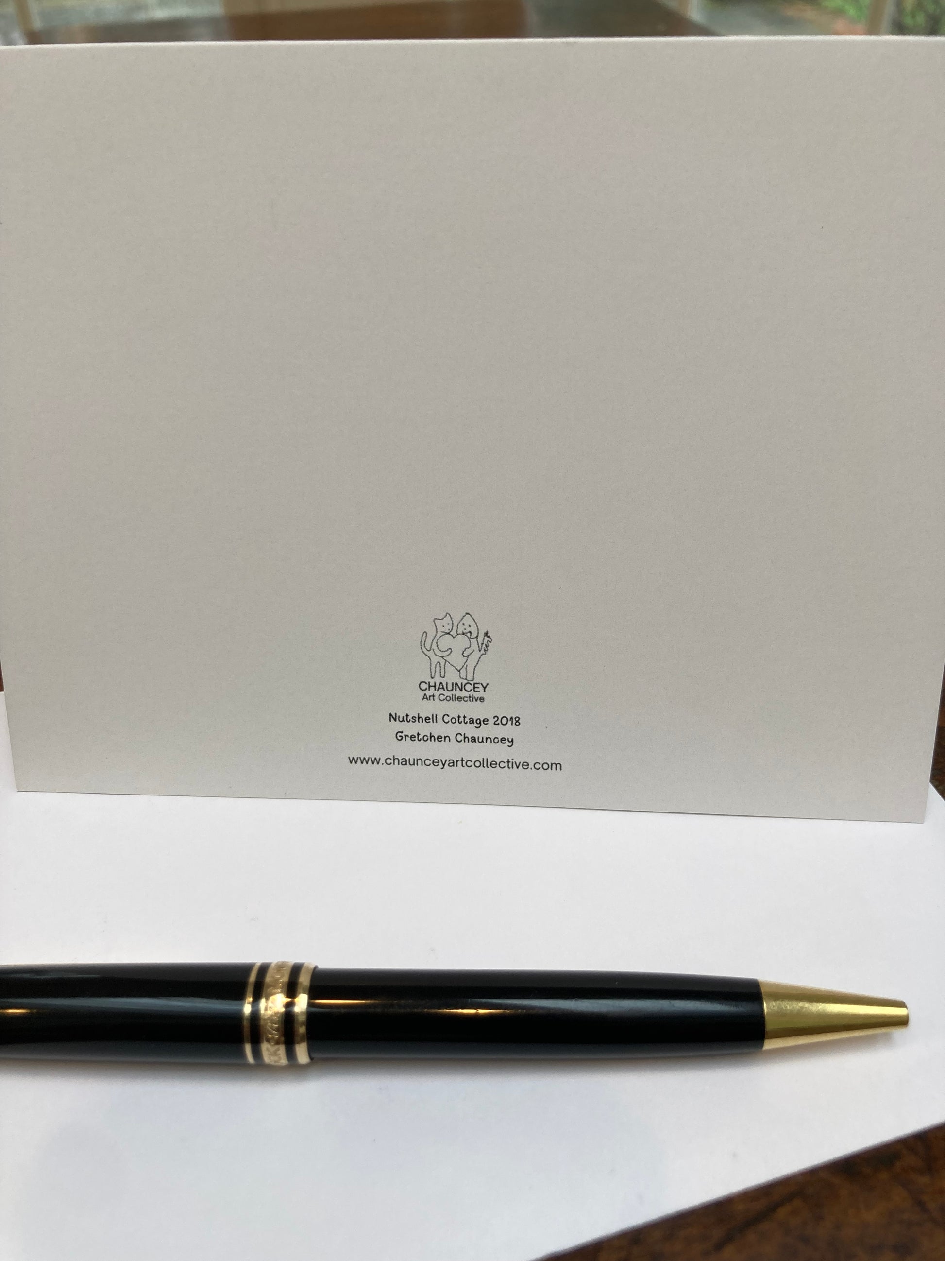 Mockup of the back of the card with the title Nutshell Cottage and the Chauncey Art Collective logo. Set on a wooden table with a fountain pen.