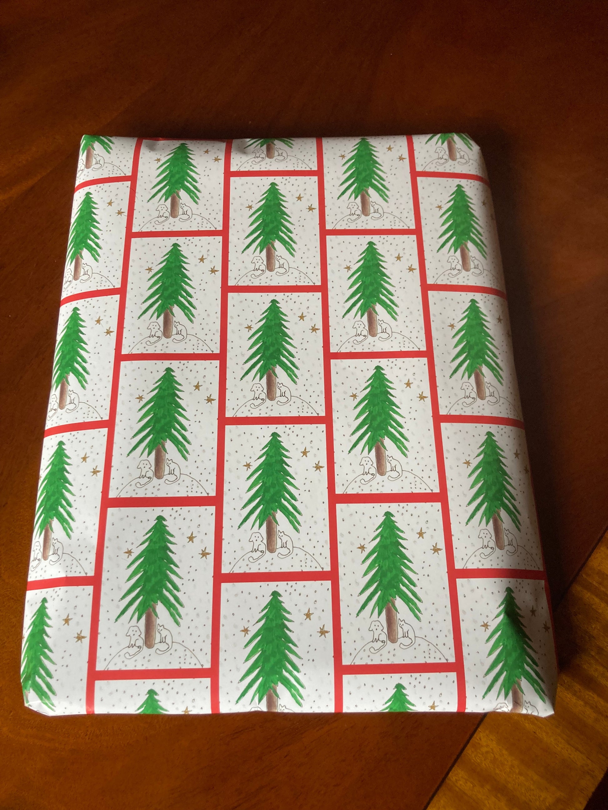 A present is wrapped in Under The Tree gift wrap highlighting the red outline to the pattern of a green pine tree with a cat and a dog under it.