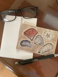 A mock up of the notecard Sea Shells By The Seashore is sitting on a wooden table with its envelope.