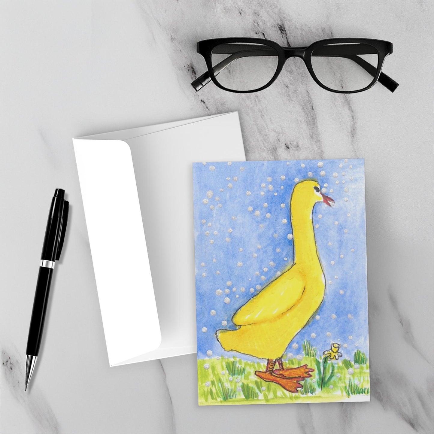 Mockup display showcasing the front of the greeting card A Few Snowflakes, Yellow Duck With Yellow Flower lying on a gray marble surface along side a white envelope, a pair of black reading glasses and a black pen.