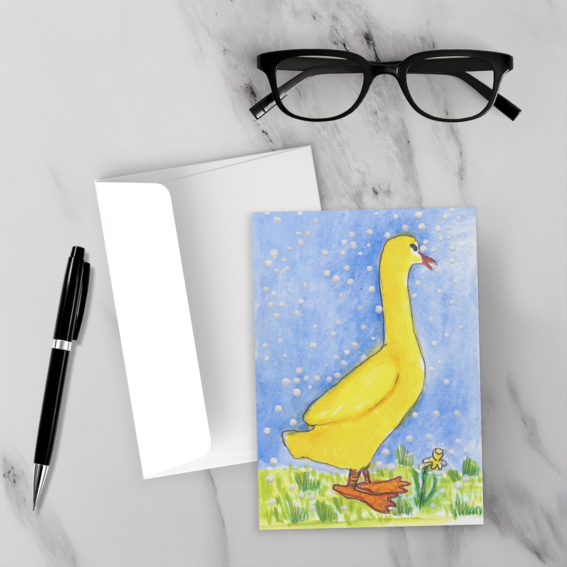 Mockup display showcasing the front of the greeting card A Few Snowflakes, Yellow Duck With Yellow Flower lying on a gray marble surface along side a white envelope, a pair of black reading glasses and a black pen.