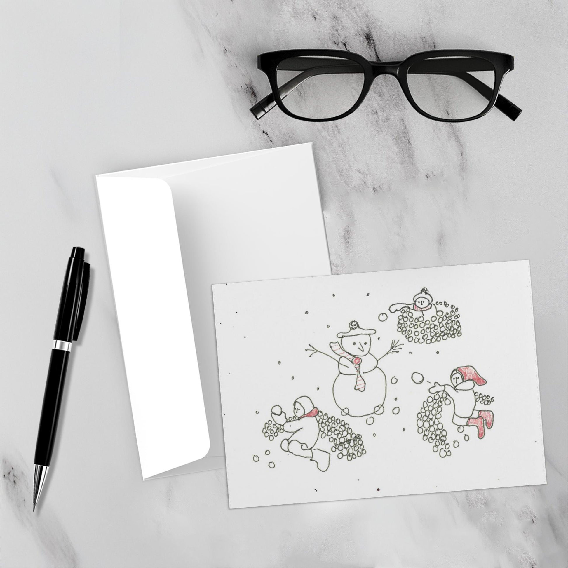 Mockup display showcasing the front of the greeting card Snowball Fight With A Snowman lying on a gray marble surface along side a white envelope, a pair of black reading glasses and a black pen.