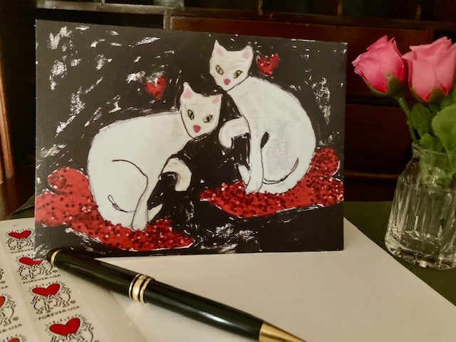 Front of the greeting card Cats On Hearts displayed on a desktop with white envelope, black pen, partial view of sheet of valentines day postage stamps and  cut glass vase with pink roses.