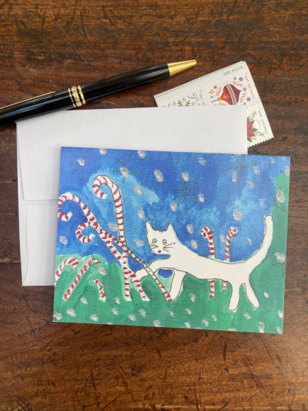 Photograph of the holiday greeting card Caspar In Candyland which is lying atop a white envelope on a brown wooden desk with some Christmas stamps in the background and a black pen