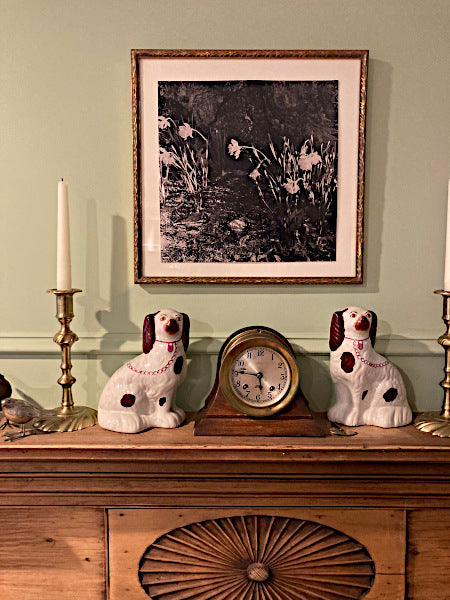 Mockup of Weston Daffodils photographic art print in a gold frame with white matting. It is hanging over a wooden mantle decorated with Staffordshire dogs, a ship clock and candlesticks.
