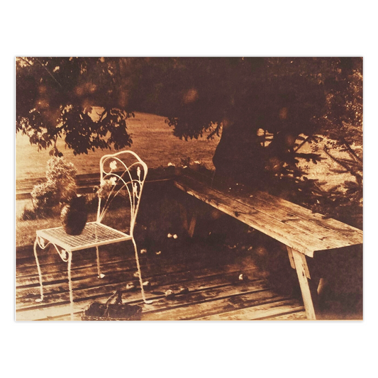 Apples On The Deck is a sepia toned photography card featuring an apple tree that overhangs a wooden deck. A wrought iron chair in white supports a vase with dried flowers.