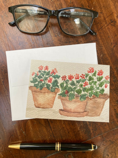 The front of Geranium Trio notecard and its envelope are displayed on a wooden table with reading glasses and a pen.