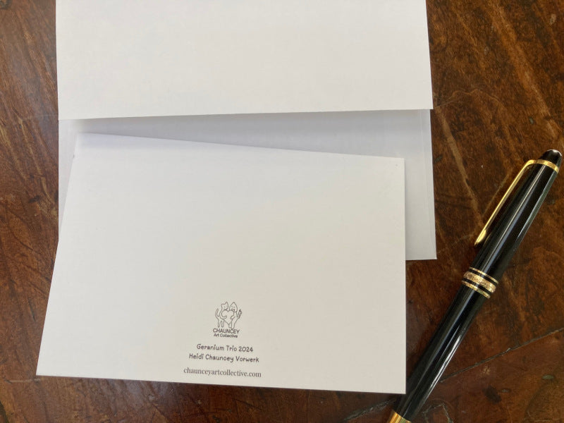 The backside of the notecard with its title, Geranium Trio and Chauncey Art Collective's logo are displayed on a wooden table with a black fountain pen.