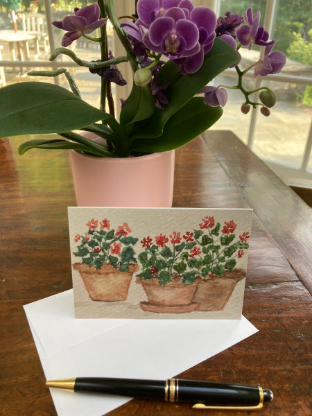 Front side of Geranium Trio notecard with its envelope are sitting on a wooden table with a blooming purple African violet and a fountain pen.