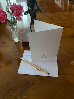 photograph of the back of the greeting card High Top Table For Daddy showing the card standing atop it's white envelope with gold pen and a vase with pink roses and an African figurine.