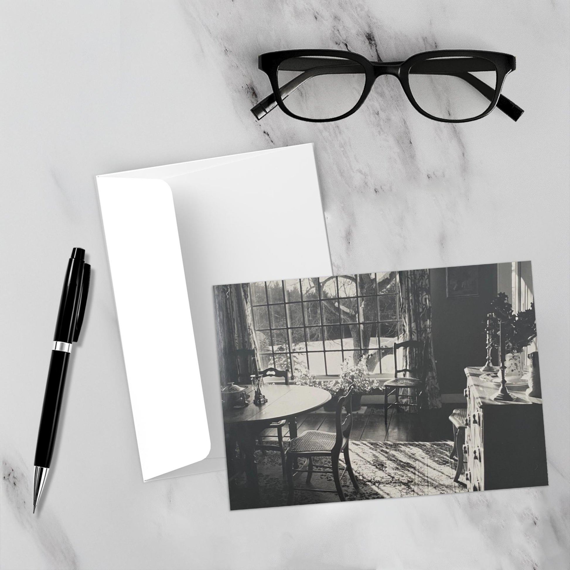 Mockup display showcasing the front of the greeting card Vermont Light lying on a gray marble surface along side a white envelope, a pair of black reading glasses and a black pen.