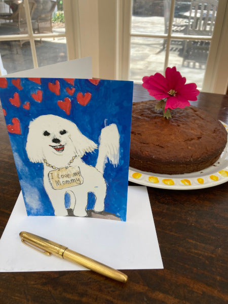A mockup of the card next to a beautiful banana cake with a flower on it. The cute image of the dog is facing the camera near the envelope and a gold fountain pen.