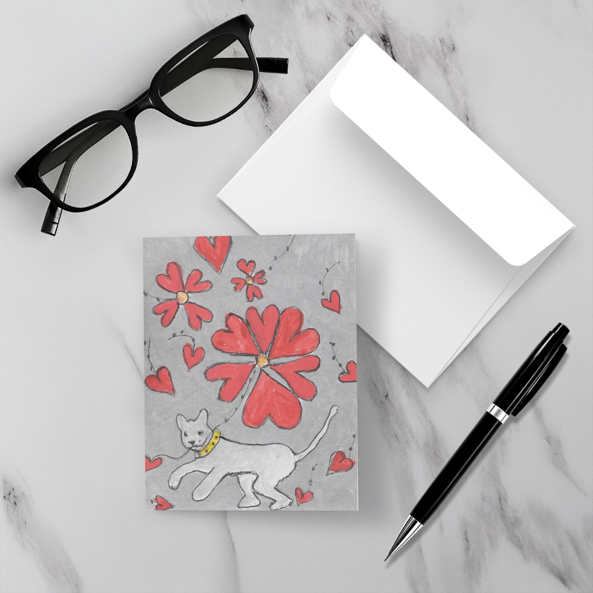 Mock up of the greeting card made just or you which displays the front of the card with the white cat holding heart balloons, a pair of black reading glasses, a black pen and a white envelope all on a gray marble surface.