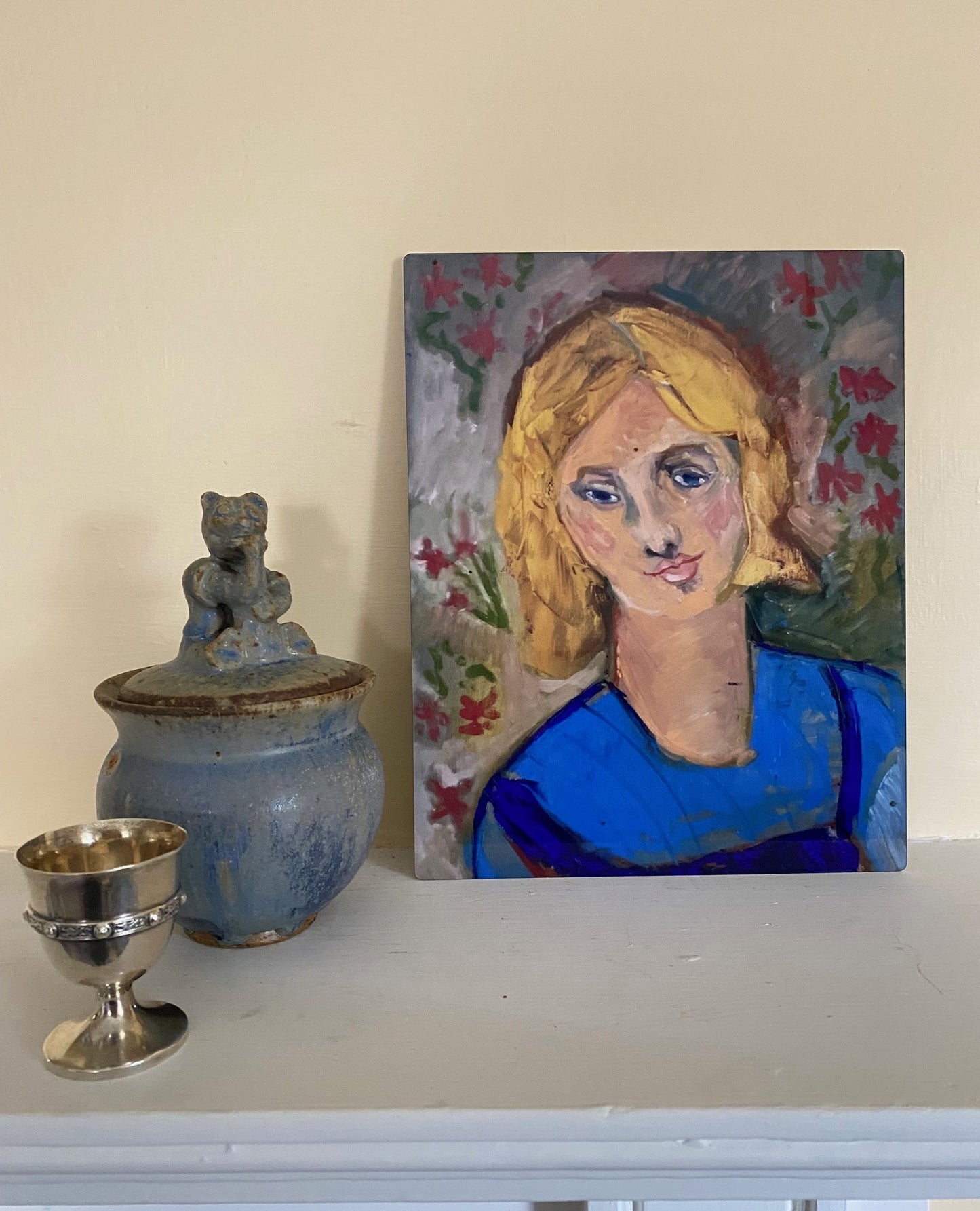 Mockup of Yellow Haired Girl metal print propped up on mantlepiece next to a blue lidded clay jar and silver egg cup.
