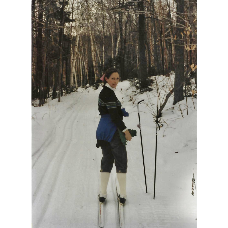 Janet Lee Chauncey on one of her favorite XC ski trails in Vermont. A woman stands on her skis with white leg warmers, gloves in hand and a pink ribbon in her hair. Inspiration for both the Trail Blazer Accessory pouch and Happy Trails flat note card  by Chauncey Art Collective.