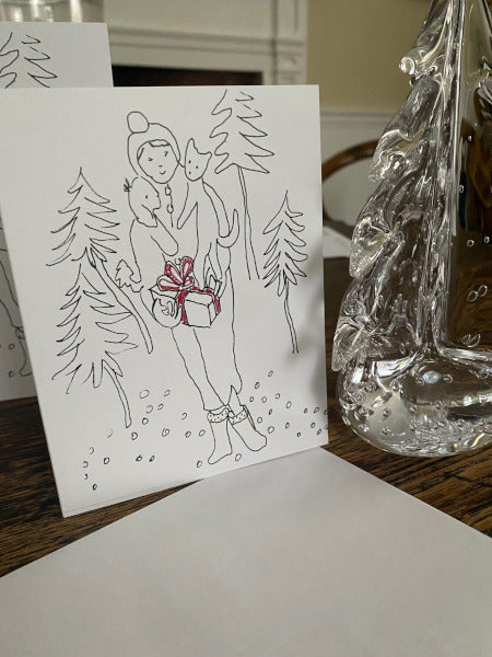 Holiday Delivery card with a person holding a gift and trees, next to a glass Christmas tree on a table.