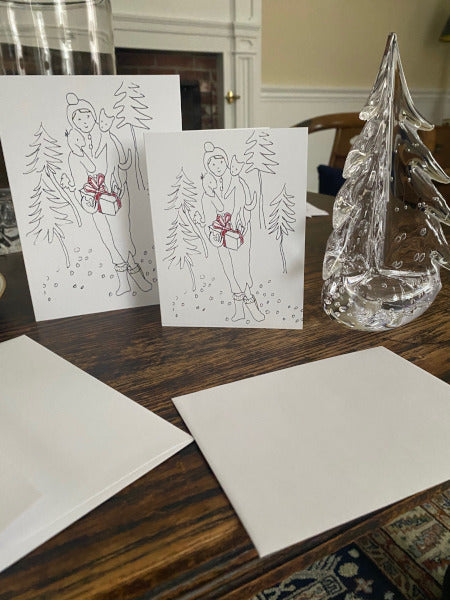 Two Christmas cards with a person and tree design on a wooden table with a glass Christmas tree.