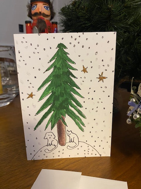 Front of the holiday greeting card Under The Tree on a wood table near a green tree.  Greeting card by Chauncey Art Collective