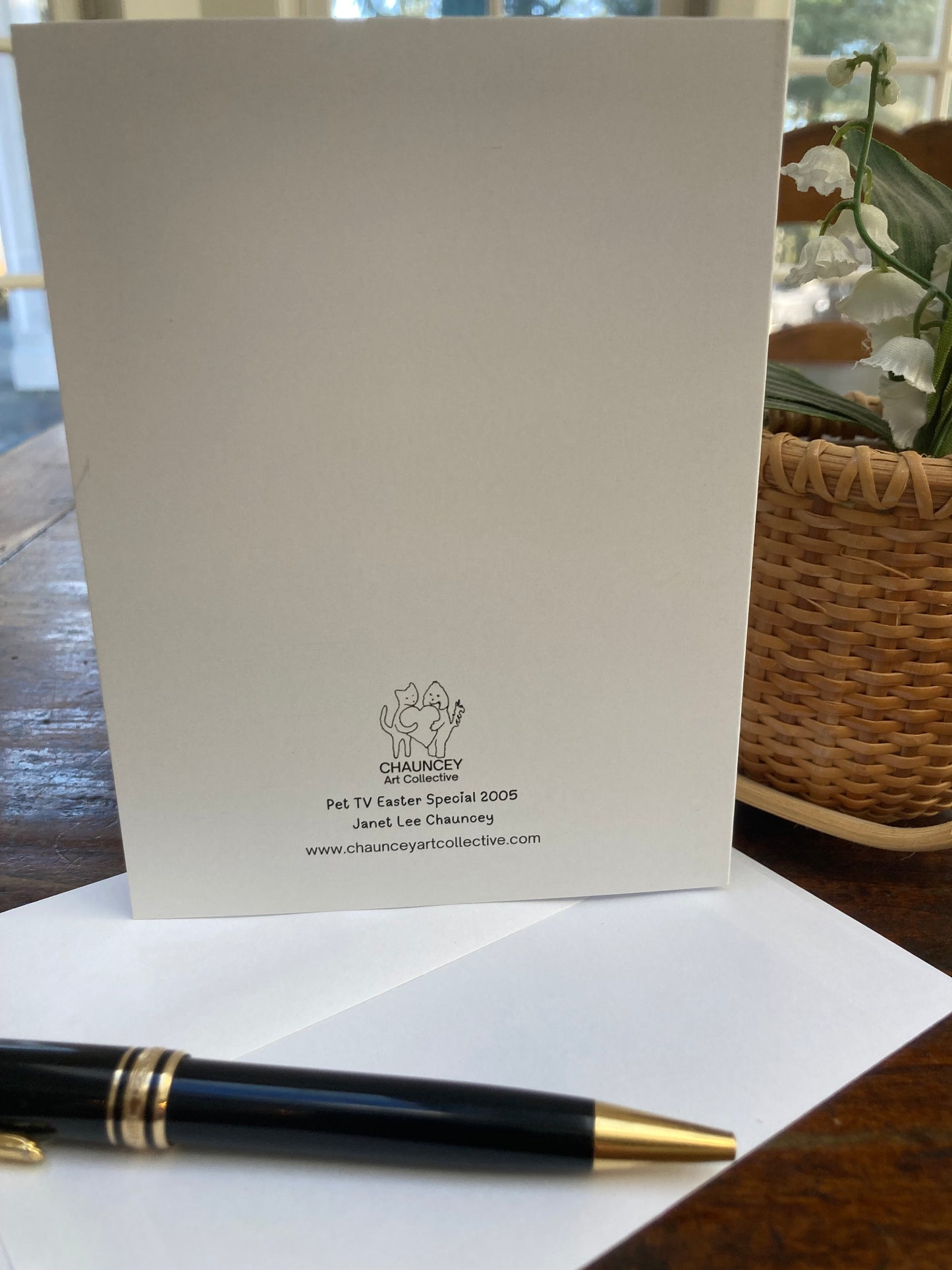 Mock up of the rear of the Easter card along with the envelope on a pen. They sit on a wooden table near a wicker backet of flowers.
