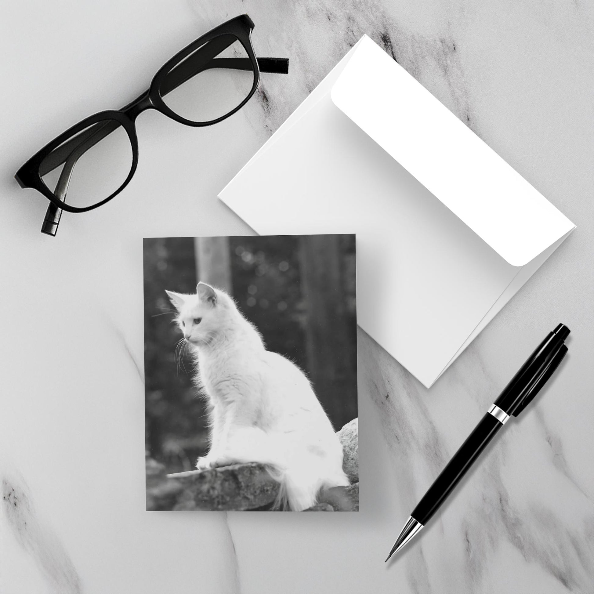 The Roxanne card is face up on a white marble backdrop along with the envelope a pen and reading glasses.