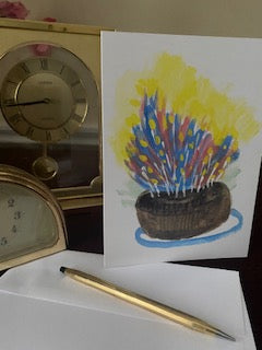 The front of the Card The Most Delicious Cake displayed on a desk with a white envelope and gold pen in the foreground and gold desk clocks visible on the left.