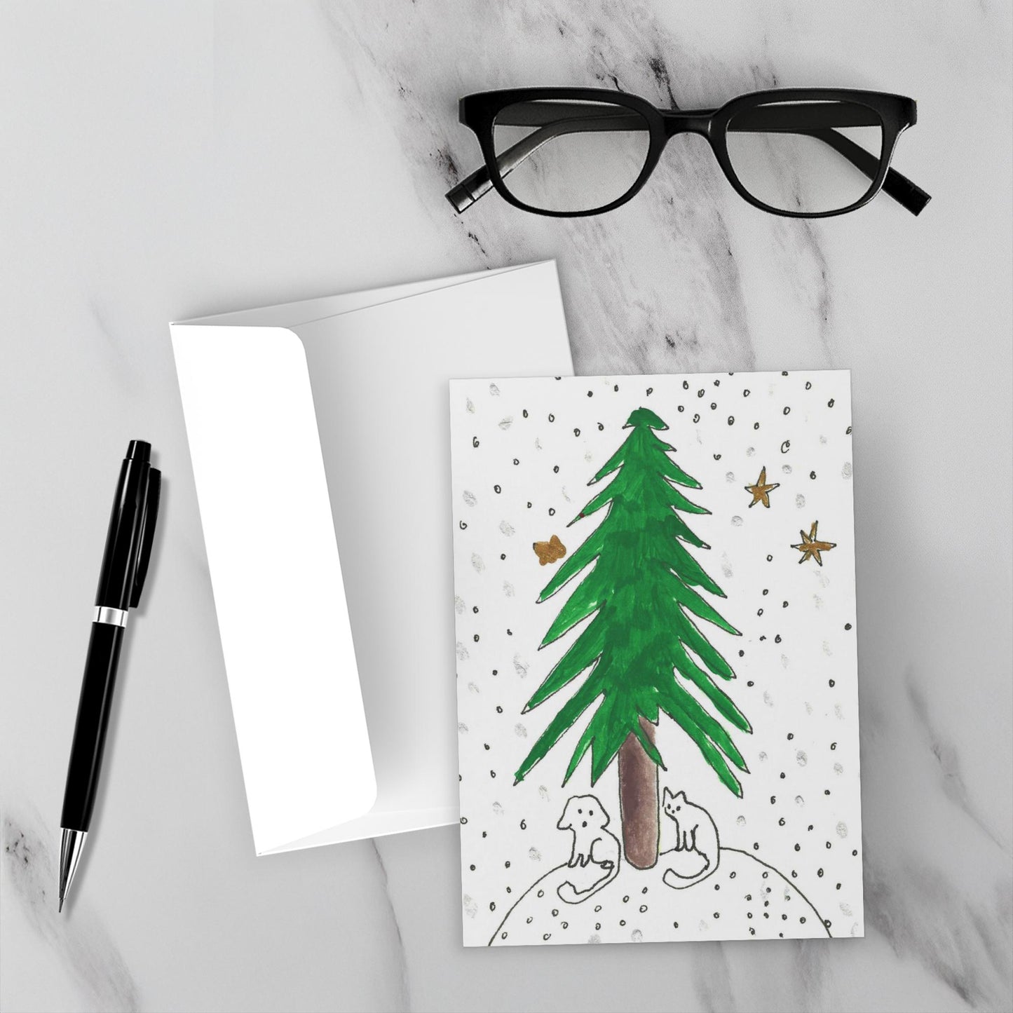 Mockup of the front of Under The Tree Christmas card and envelope on a white marble background with black rimmed glasses and a black pen.