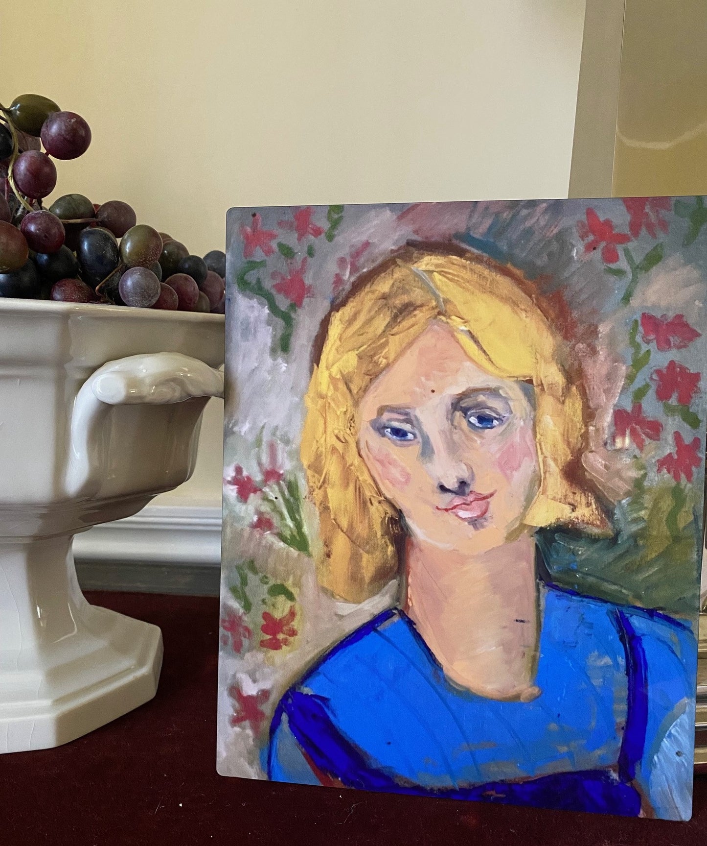 Mockup of Yellow Haired Girl metal print resting on red velvet desk top next to a white ceramic bowl filled with red grapes.