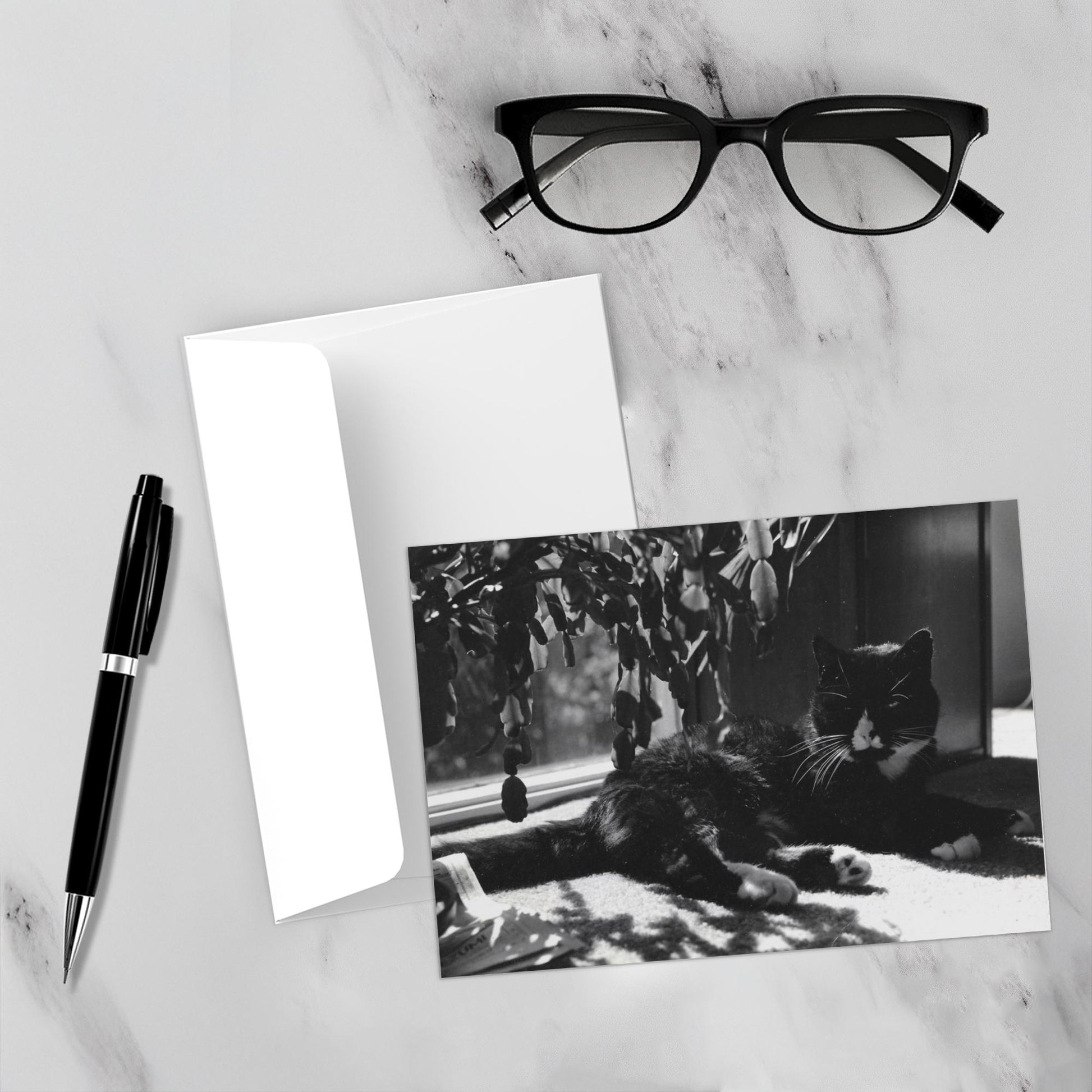 Mockup display showcasing the front of the greeting card Puddy Tat In The Window lying on a gray marble surface along side a white envelope, a pair of black reading glasses and a black pen.
